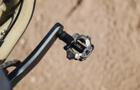 Primer plano de un pedal de bicicleta Shimano DEORE XT con sistema SPD fijado a un brazo de biela, con una superficie de tierra arenosa al fondo, lo que resalta la perfecta configuración de los pedales para senderos.