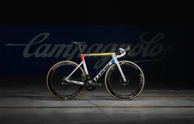 Una bicicleta de carretera blanca y azul con detalles en amarillo se destaca sobre un fondo oscuro con la palabra "Campagnolo" en la parte posterior.