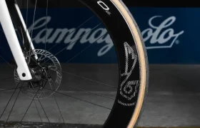 Primer plano de la rueda delantera de una bicicleta con llanta y neumático negros con el logotipo de Campagnolo. En el fondo se ve un cartel de Campagnolo borroso sobre una pared azul.