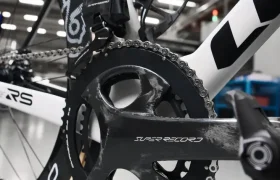 Primer plano de la transmisión de una bicicleta, con un desviador trasero, un plato grande y un pedal, sobre un fondo interior.