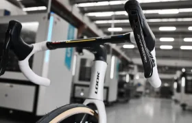 Primer plano de una bicicleta de carreras en un taller, con el manillar y el sillín en primer plano. Al fondo se ven máquinas y equipos industriales.