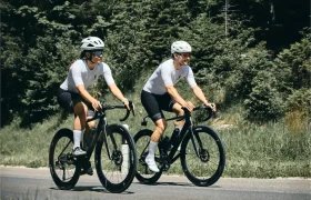 Dos ciclistas con casco y ropa de ciclismo blanca a juego montan en bicicletas de carretera HADRON³ Ultimate 650 por una carretera asfaltada bordeada de hierba y árboles frondosos, empujando el límite UCI con aerodinámica superior.