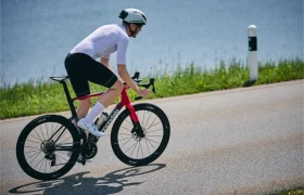 Un ciclista con maillot y casco blancos monta en una bicicleta de carretera roja y negra cerca del agua y la hierba, haciendo gala de aerodinámica con la Side suiza HADRON³ Ultimate 650 mientras se mantiene dentro del límite UCI.