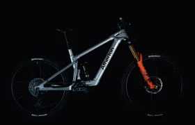 La Avinox M2S es una bicicleta de montaña de suspensión total con cuadro plateado, ruedas negras y horquilla delantera naranja, que se muestra sobre un fondo oscuro.