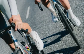 Dos ciclistas, con zapatillas y calcetines blancos, circulan en bicicleta de carretera por una calle asfaltada bañada por el sol; solo se ven la parte inferior de sus cuerpos y algunos detalles de las bicicletas.