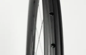 Primer plano de una llanta de bicicleta negra con los agujeros de los radios visibles, vista de perfil sobre un fondo blanco liso.