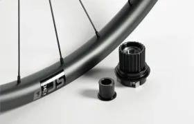 Un primer plano de la llanta de una bicicleta con radios, junto a dos componentes metálicos negros del buje, sobre un fondo blanco.