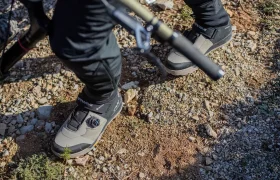 Una persona que lleva calzado de montaña gris y negro está de pie sobre un terreno rocoso, con una caña de pescar en la mano; solo se le ven las pantorrillas y los pies.