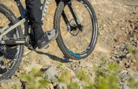 Una persona montando en bicicleta de montaña por un sendero rocoso, en la que se aprecian las ruedas y los pedales de la bicicleta, así como la parte inferior de las piernas del ciclista, que lleva ropa negra.