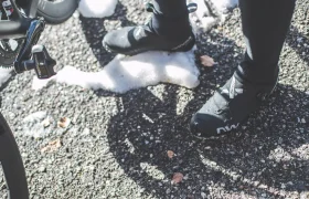 Primer plano de los pies de un ciclista, calzado con zapatillas de ciclismo negras, junto a una bicicleta sobre una superficie de asfalto parcialmente cubierta de nieve.