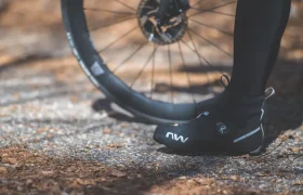 Una persona que lleva zapatillas de ciclismo negras está de pie junto a una rueda de bicicleta en un sendero iluminado por el sol y cubierto de hojas.