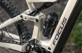 Primer plano del cuadro blanco de una bicicleta de montaña eléctrica FOCUS e-MTB, en el que se aprecian el pedal, la cadena, el bidón y la suspensión trasera sobre un camino de grava, lo que refleja a la perfección el diseño elegante que caracteriza a la serie JAM.