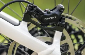 Primer plano de una pinza de freno de disco Tektro montada en el cuadro blanco de la bicicleta eléctrica de montaña JAM 2027, con el disco de freno visible y los radios al fondo.