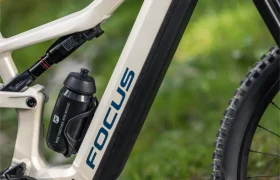 Primer plano del cuadro de una bicicleta de montaña eléctrica Focus JAM de color beige, con una batería negra, un bidón y una vista parcial de las ruedas delanteras y traseras; césped y vegetación difuminada al fondo, lo que pone de relieve el diseño avanzado de la Gama FOCUS e-MTB 2027.