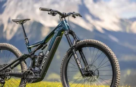 Una bicicleta de montaña eléctrica FOCUS de color negro, con ruedas grandes, está aparcada sobre la hierba, con las montañas y un fondo natural difuminado.