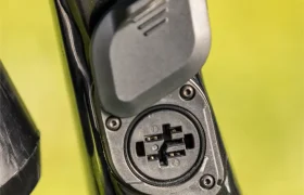Primer plano de un puerto de recarga de una bicicleta eléctrica SAM con la tapa protectora parcialmente abierta, montado en un cuadro Gama FOCUS e-MTB 2027 de color negro brillante, sobre un fondo verde difuminado.
