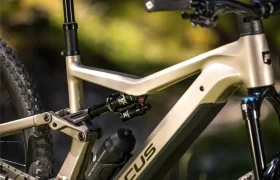 Primer plano de una bicicleta de montaña Focus JAM de doble suspensión de color beige con un bidón de agua fijado al cuadro, fotografiada al aire libre bajo la luz del sol.