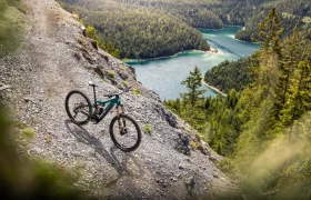 Una bicicleta de montaña FOCUS e-MTB 2027 descansa sobre una ladera rocosa con vistas a un valle boscoso y a un sinuoso lago azul que se extiende más abajo.