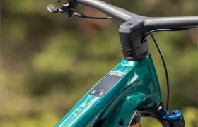 Primer plano del manillar, la pantalla y la suspensión delantera de una bicicleta eléctrica de montaña JAM 2027 de color verde azulado, con un bosque desenfocado al fondo.