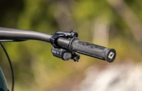 Primer plano del manillar de una bicicleta FOCUS e-MTB 2027, con puños de goma texturizados y botones de control electrónico incorporados, sobre un fondo difuminado de un paisaje al aire libre.