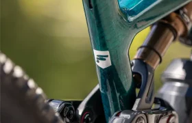 Primer plano de un cuadro de bicicleta de montaña JAM de color verde azulado, en el que se aprecian la suspensión trasera, la tija del sillín y parte de la rueda trasera, con un fondo verde difuminado.
