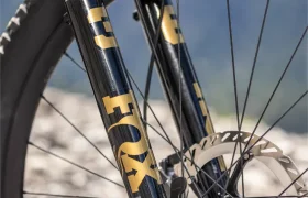 Primer plano de una horquilla de suspensión delantera de una bicicleta de montaña con el logotipo «FOX» en dorado y negro, un disco de freno y un fondo rocoso, que refleja a la perfección el rendimiento resistente que cabría esperar de una e-MTB de FOCUS como la JAM o la SAM.