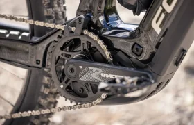 Primer plano del juego de bielas, el plato y el pedal de una bicicleta de montaña. La cadena está colocada y el cuadro lleva la marca THRON con un acabado negro mate. Se ven tierra y piedras en el suelo, lo que sugiere que podría tratarse de un modelo de e-MTB de 2027 listo para los senderos.