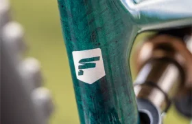 Primer plano del cuadro de una bicicleta eléctrica de montaña Gama FOCUS 2027 de color verde azulado, con un logotipo blanco en forma de escudo, en el que se aprecia la textura detallada y parte de la suspensión trasera.
