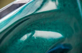 Primer plano de una superficie curvada, de color turquesa y brillante, con detalles texturizados —posiblemente parte de una bicicleta eléctrica de montaña FOCUS— y una vista parcial de un objeto rectangular en la parte superior izquierda.