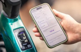 Una mano sostiene un smartphone en el que se ven los ajustes de una bicicleta eléctrica; en la pantalla aparece 750 W y «e-MTB 2027» resaltado; al fondo se ve un panel de control SAM.