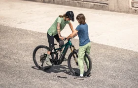 Dos personas están de pie en la acera junto a una bicicleta e-MTB 2027; una de ellas, JAM, está sentada en el sillín, mientras que la otra, SAM, sujeta el manillar para apoyarse.