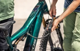 Un ciclista utiliza un manómetro digital para comprobar o inflar la presión de aire del amortiguador trasero de una bicicleta eléctrica de montaña FOCUS e-MTB 2027, mientras otra persona —que se ve parcialmente— se encuentra de pie cerca de él.