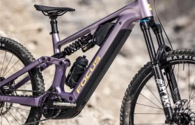 Una bicicleta de montaña eléctrica de doble suspensión Focus JAM de color morado, perteneciente a la colección Gama FOCUS e-MTB 2027, equipada con suspensión Fox y neumáticos Maxxis, está aparcada en un terreno rocoso.