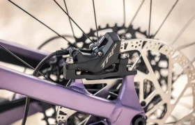Primer plano de una pinza de freno de disco de bicicleta montada en un cuadro de bicicleta eléctrica de montaña (e-MTB) de color morado, con el disco de freno y los radios de la rueda trasera visibles al fondo.