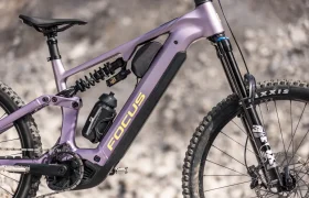 Primer plano lateral de una bicicleta eléctrica de montaña (e-MTB) Focus JAM de 2027 con suspensión total y color morado, en el que se aprecian el cuadro, los amortiguadores, el portabidones y parte de las ruedas delantera y trasera sobre un fondo difuminado.