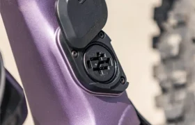 Primer plano del puerto de carga de una bicicleta eléctrica de montaña (e-MTB) FOCUS con una tapa protectora en un cuadro de color morado, junto a la rueda trasera y al pivote de la suspensión.