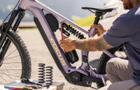Una persona está ajustando el muelle de la suspensión trasera de una bicicleta eléctrica de montaña FOCUS e-MTB de color morado; junto a ella, en el suelo, hay herramientas y muelles de repuesto.
