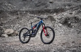 Una bicicleta de montaña eléctrica Gama FOCUS e-MTB 2027, de color azul y rojo, con suspensión total, se encuentra sobre un terreno rocoso con un fondo gris neutro.