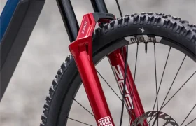 Primer plano de la rueda delantera de una bicicleta eléctrica de montaña (e-MTB) FOCUS 2027, con una horquilla de suspensión RockShox roja y un freno de disco, sobre un fondo gris difuminado.
