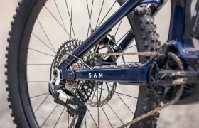 Primer plano del desviador trasero, los piñones y la cadena de una bicicleta de montaña eléctrica Gama FOCUS 2027 de color azul oscuro, con el cuadro marcado con la inscripción «SAM», en el que se aprecian los dibujos del dibujo de los neumáticos.