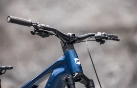 Primer plano del manillar y la parte delantera del cuadro de una bicicleta eléctrica de montaña (e-MTB) azul FOCUS 2027, en el que se ven los cables de freno y el fondo gris aparece difuminado.