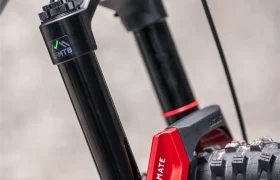 Primer plano de una horquilla de suspensión delantera de una bicicleta eléctrica de montaña (e-MTB) de la marca FOCUS, en el que se aprecian los montantes negros, la parte inferior de la horquilla en rojo con la inscripción «ULTIMATE» y una parte del neumático delantero sobre un fondo gris.