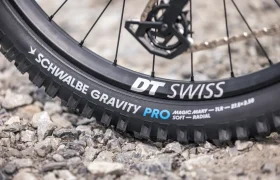 Primer plano de una rueda de bicicleta de montaña con llanta «DT Swiss» y neumático «Schwalbe Gravity Pro», montada en una e-MTB de la marca JAM, que se muestra apoyada sobre una superficie de grava.