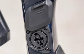 Primer plano del cuadro de una bicicleta eléctrica de montaña (e-MTB) de la marca FOCUS, en el que se aprecia el puerto de carga y su tapa, con los tornillos y los componentes de la suspensión visibles a su alrededor.