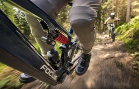 Primer plano de una bicicleta eléctrica de montaña Gama FOCUS 2027 en movimiento por un sendero forestal, con otro ciclista siguiéndola por detrás.