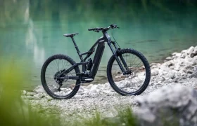 Una bicicleta eléctrica de montaña FOCUS 2027 de color negro, con neumáticos gruesos, está aparcada en un sendero rocoso junto a una masa de agua de color turquesa, con vegetación difuminada en primer plano.