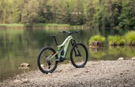 Una bicicleta de montaña eléctrica FOCUS de color verde está aparcada en una orilla rocosa junto a un lago en calma, con el bosque y la vegetación visibles al fondo.