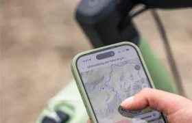 Persona con un smartphone en el que se ve un mapa de rutas ciclistas, junto al manillar de una bicicleta eléctrica de montaña JAM 2027 de color verde, al aire libre.