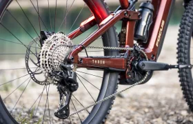 Primer plano de la rueda trasera y el sistema de cambios de una bicicleta de montaña Focus Thron roja, en el que se aprecian el desviador, el cassette, la cadena y parte del cuadro —similar a lo que se puede encontrar en modelos de e-MTB de FOCUS como la JAM o la SAM— con grava en el suelo.