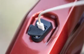 Primer plano de un cable de carga blanco enchufado al puerto de carga del cuadro de una bicicleta eléctrica de montaña (e-MTB) FOCUS 2027 de color rojo metalizado.